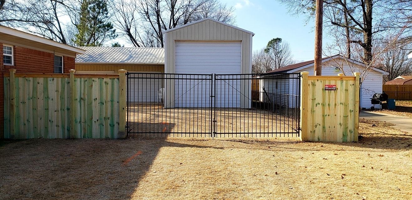 Wooden fenced driveway with open gate in front of a small white shed and trees
