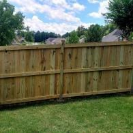 Wooden backyard fence with a grassy lawn and trees under a cloudy sky