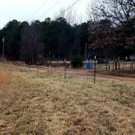 Grassy field with a wire fence, trees in the background, and a blue sign near a dirt path