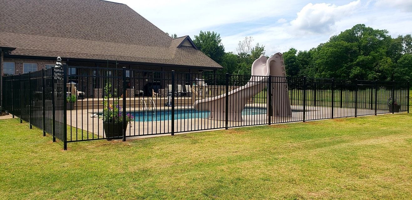 Backyard pool behind black metal fence beside a brick house and lawn
