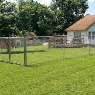 Chain-link fence around a green lawn beside a house with trees in the background
