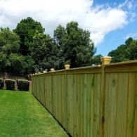 Wooden privacy fence beside a green lawn and trees under a partly cloudy sky