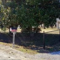 Yard with a tree, fence, and a small sign by a gravel roadside.