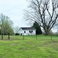 White house with black shutters in a grassy yard, framed by leafless trees and fence posts under a cloudy sky
