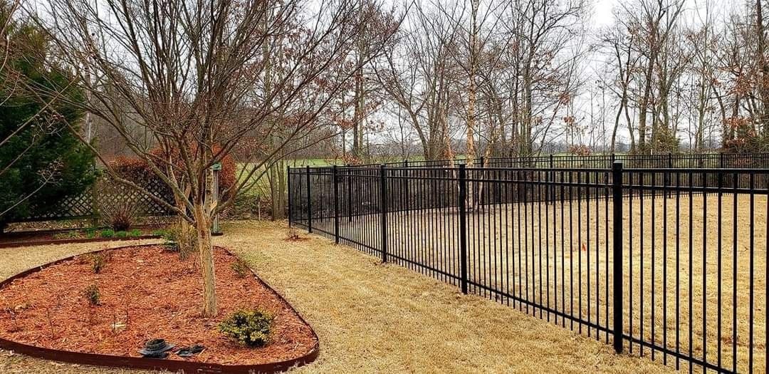 Curved mulched garden bed beside a black metal fence along a grassy path in winter trees