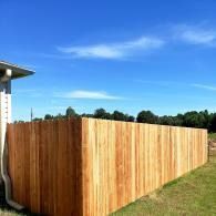 Wooden privacy fence with a sloped top beside a house and grassy yard under a blue sky
