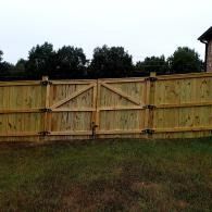 Wooden backyard fence with a double gate in front of trees and grass under an overcast sky