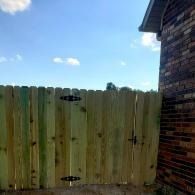 Weathered green wooden fence beside a brick house under a blue sky.