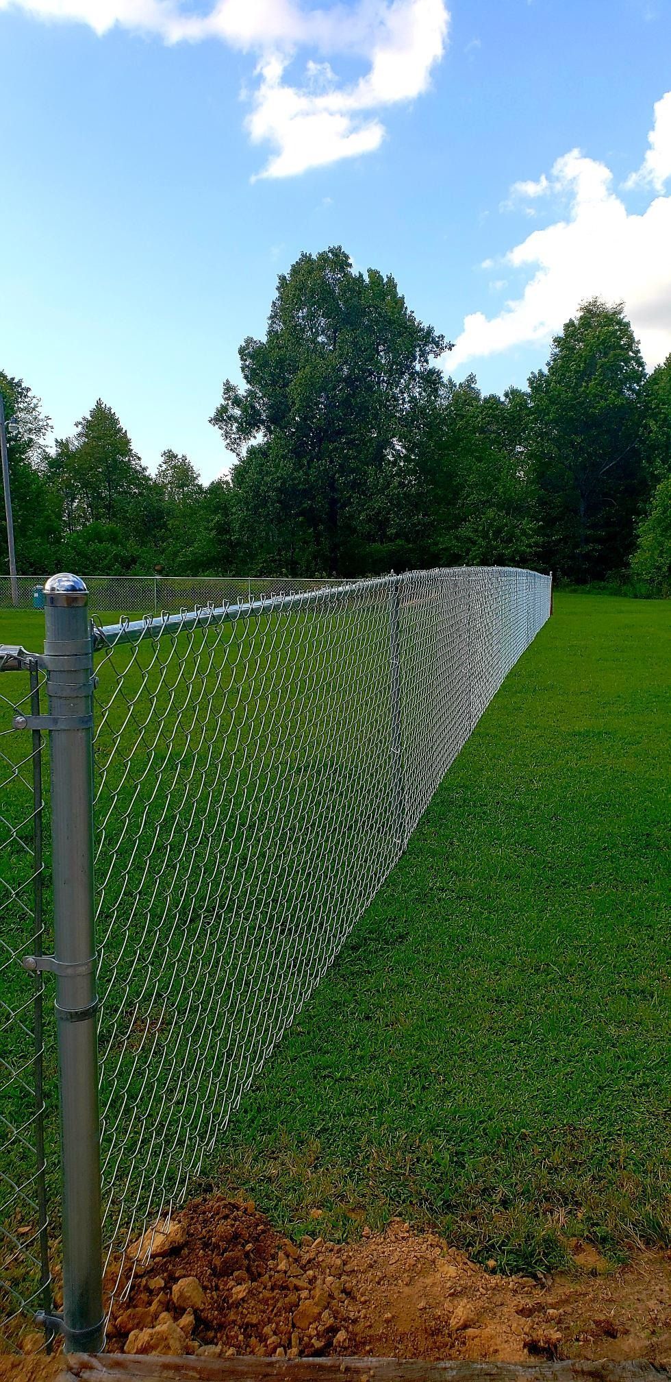 Chain-link fence curving along a green lawn beside trees under a blue sky with clouds