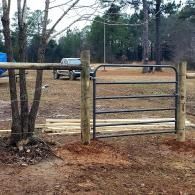 Metal farm gate opening onto a dirt driveway with a parked truck and trees in the background
