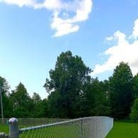 Fence curving along a green field beside trees under a bright blue sky with clouds