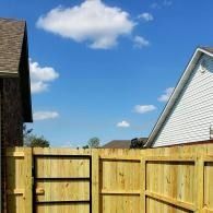 Wooden backyard fence with open gate under a blue sky with clouds, framed by house rooftops.