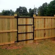 Wooden privacy fence with a small black gate in a grassy yard, trees visible behind it.