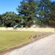 Grassy field beside a paved path, with a wooden fence and large trees under a clear blue sky
