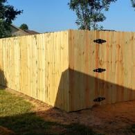 Wooden fence corner in a grassy yard with black hinges on the right panel, lit by afternoon sun.
