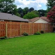 Wooden privacy fence beside a green lawn with trees and houses under a cloudy sky