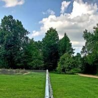 Green grassy field with a long narrow fence leading to dense trees under a cloudy blue sky