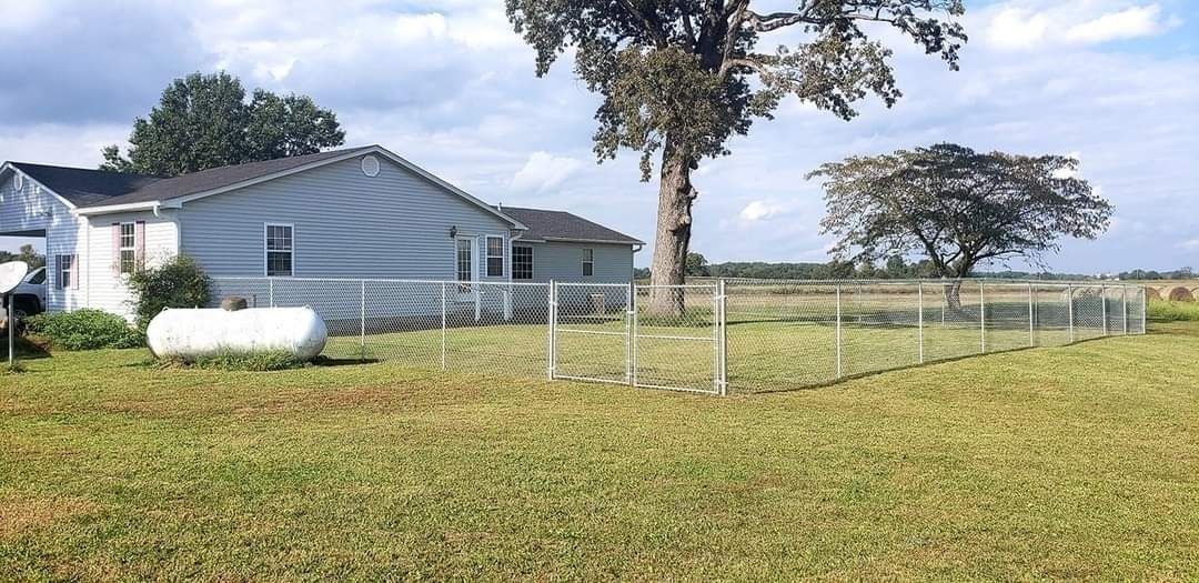 Small white house with fenced yard and grassy field under a clear sky.