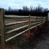 Wooden fence beside a grassy field under a cloudy sky, with leafless trees in the background.