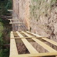 Wooden boardwalk under construction along a dirt hillside trail, with several unfinished sections.