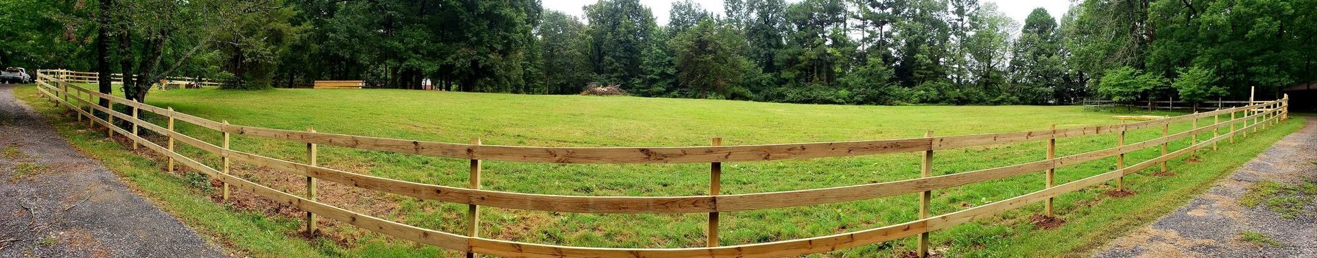 Wooden fence bordering a green grassy field with trees under a cloudy sky