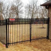 Black metal fence and gate enclosing a yard with bare trees and a brick building.