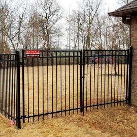 Black metal fence enclosing a dry yard beside a building, with bare trees in the background