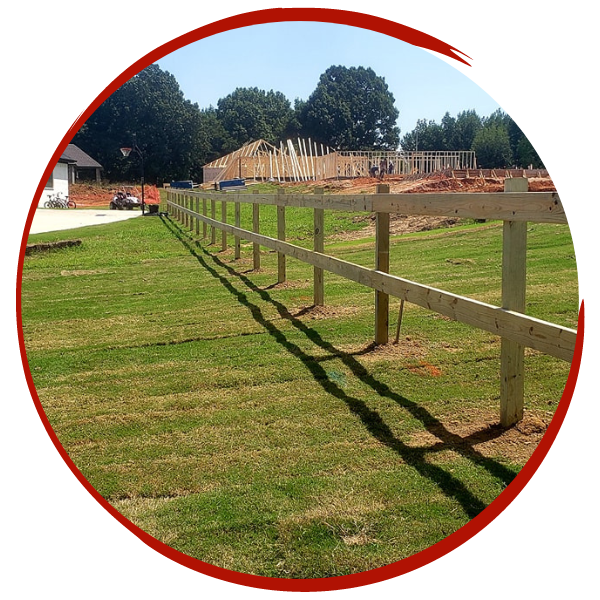 Red fence posts and rails along a mowed grassy field with trees and buildings in the background