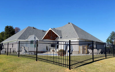 Single-story house with gray roof behind a black metal fence on a grassy lot under a blue sky