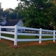 White ranch fence in a grassy yard with trees and houses in the background.