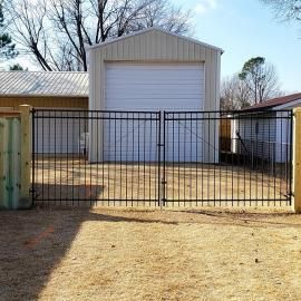 Black metal gate in front of a white garage or storage building on a dirt driveway.