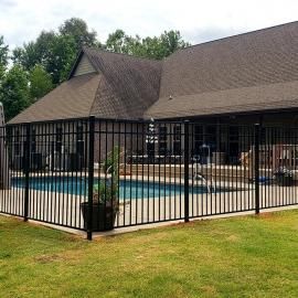 Backyard swimming pool enclosed by a black metal safety fence beside a house.
