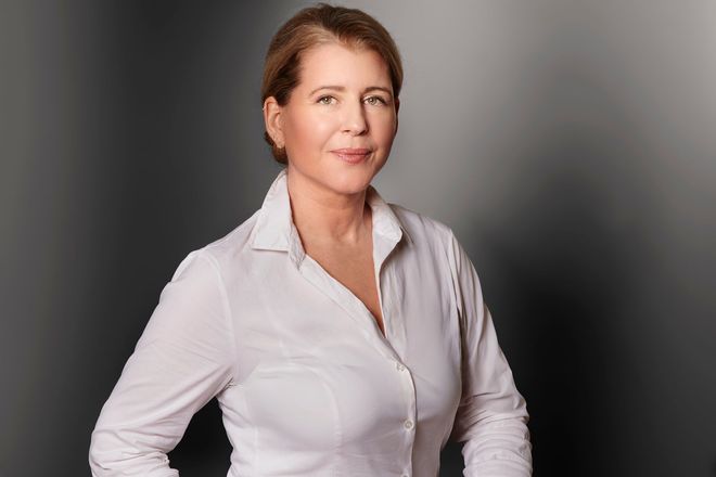 Eine Frau in einem weißen Hemd steht mit verschränkten Armen vor einem Fenster.