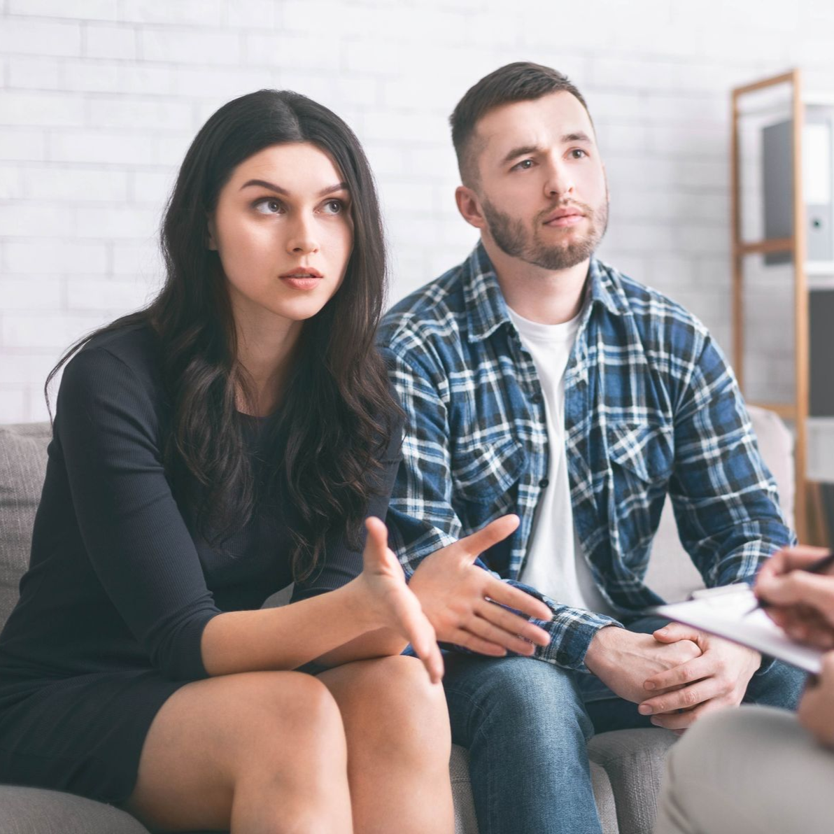 Therapist in blue chair talking to a couple on a sofa. They appear serious.