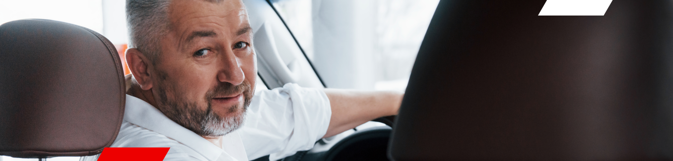 Um homem de camisa branca sorri enquanto está sentado em um carro, olhando para trás.