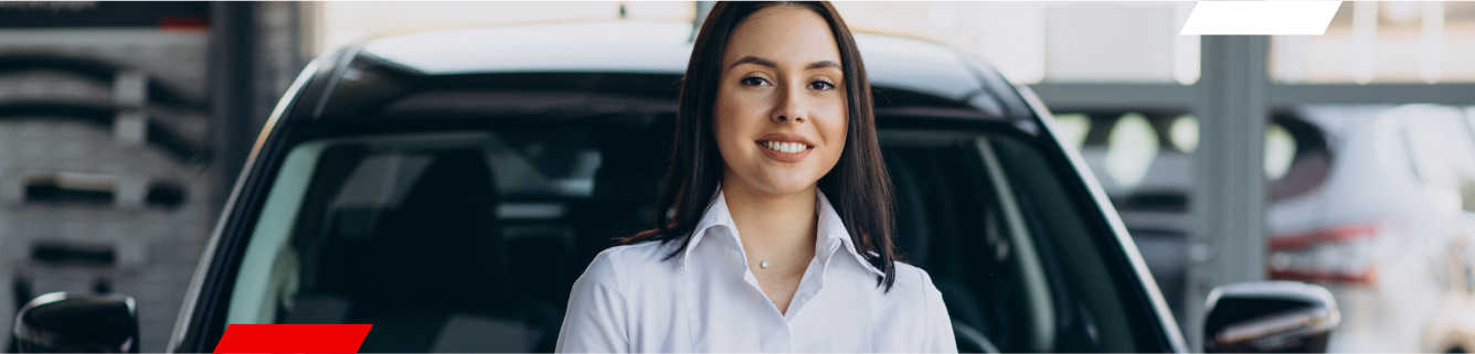 Mulher de camisa branca sorrindo, em pé na frente de um carro preto em uma concessionária de veículos.