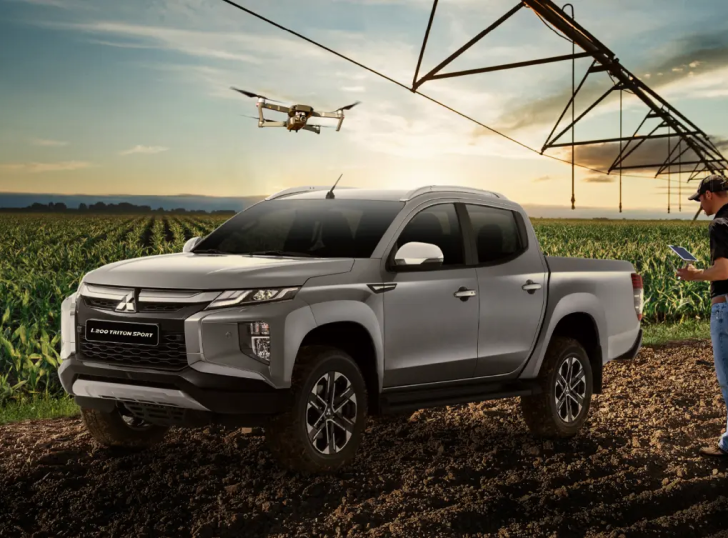 Caminhonete Mitsubishi prateada em um campo, drone sobrevoando, pessoa operando um tablet.