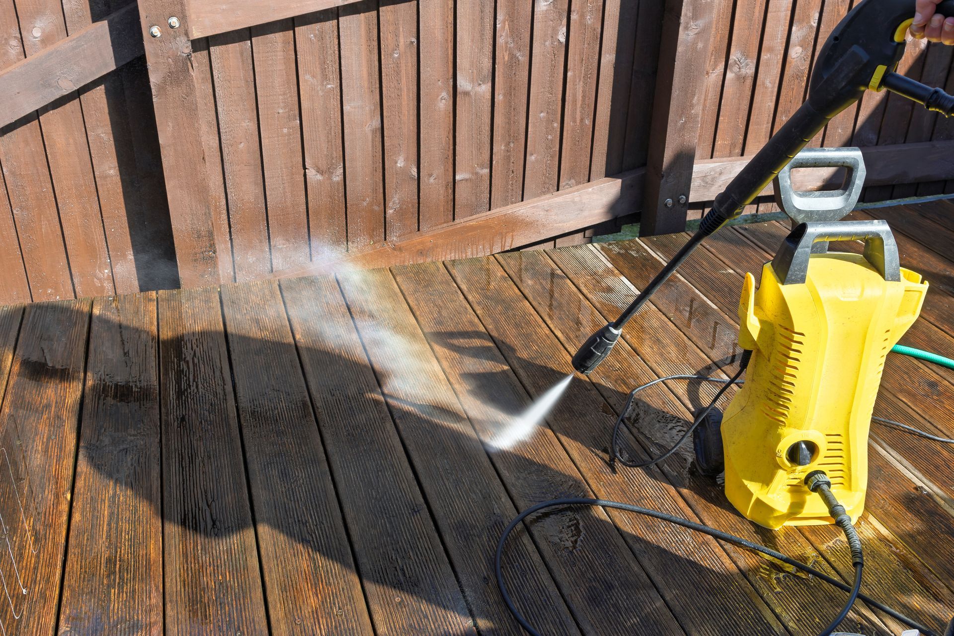 Yellow pressure washer spraying water on a wooden deck beside a fence