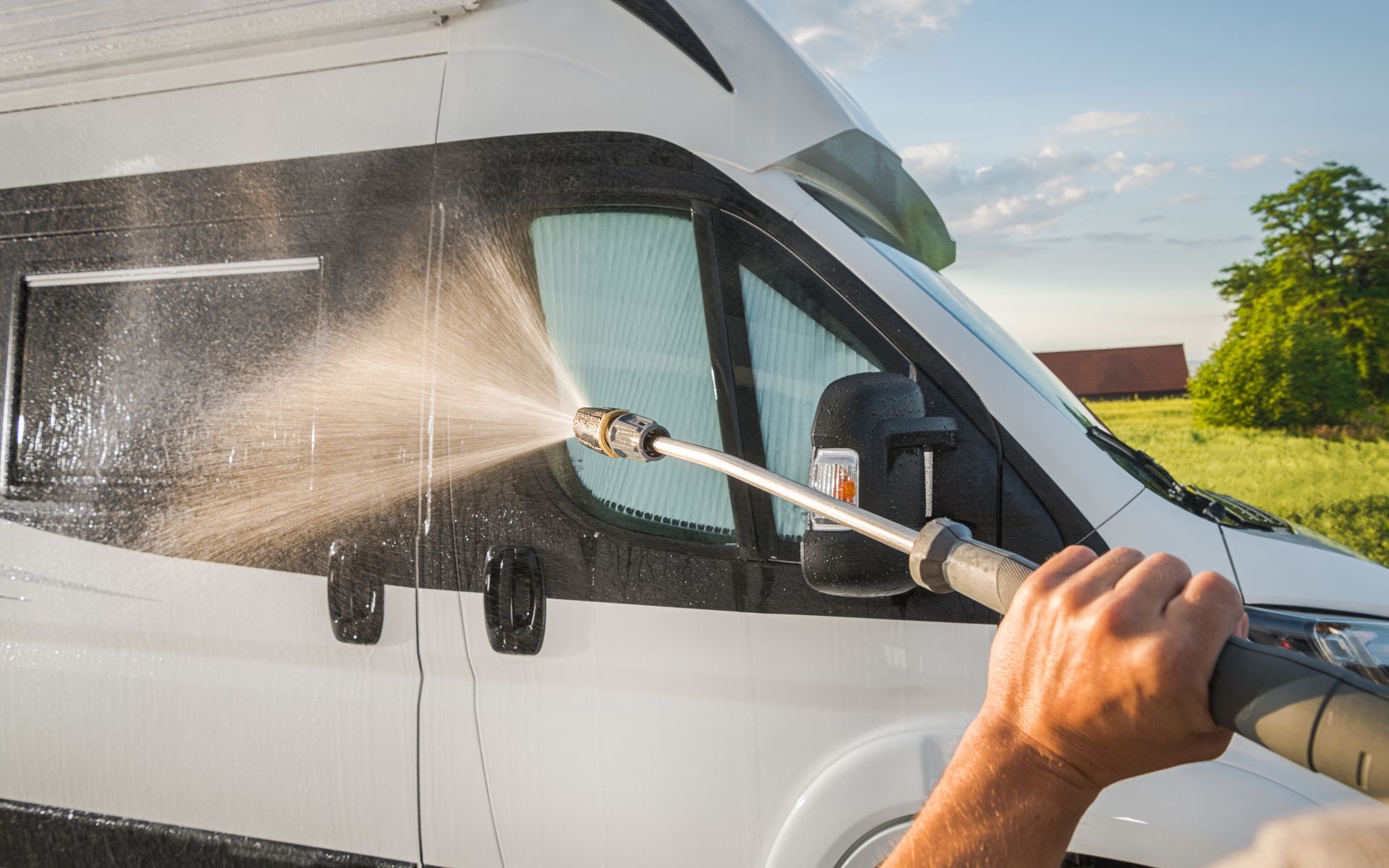 Person pressure-washing a white van with a hose outdoors on a sunny day
