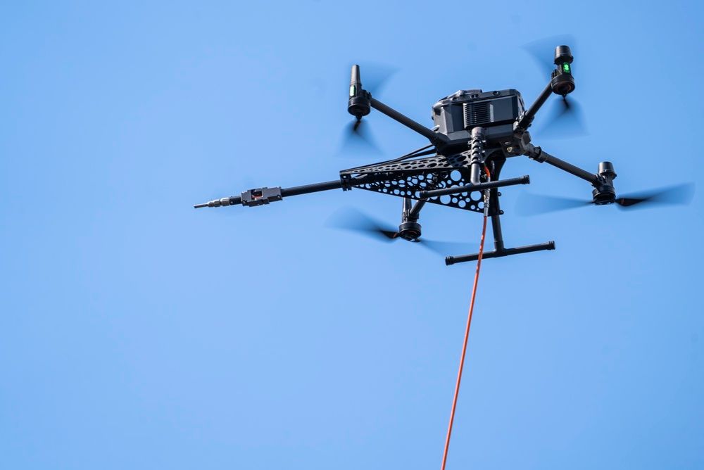 Black quadcopter drone with red cord flying against a clear blue sky