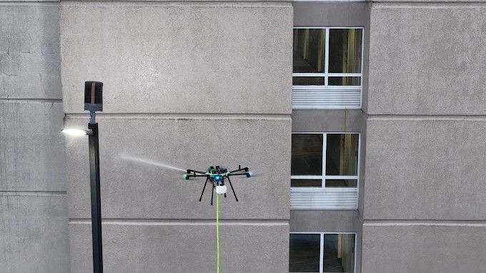 Drone hovering beside a gray building wall with a green tether