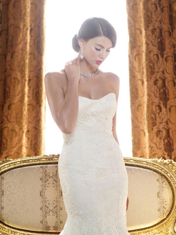 Woman in a strapless white dress standing before a gold-trimmed sofa and patterned curtains, looking down.