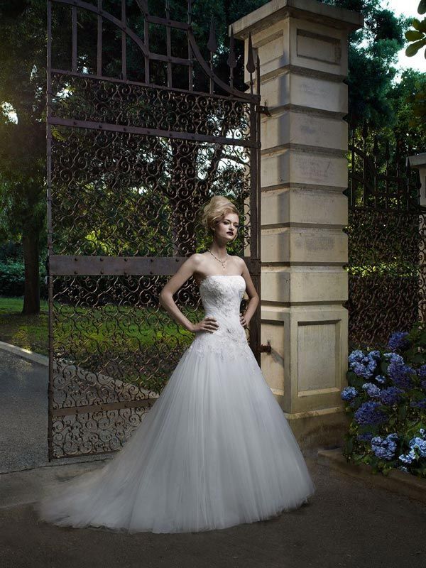 Woman in a white strapless gown standing by a wrought-iron gate and stone pillar at dusk.
