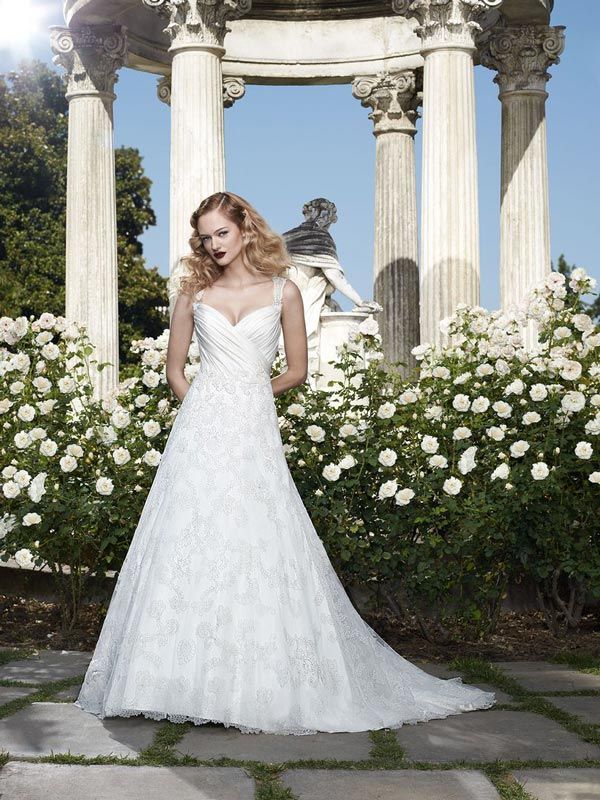 Bride in a white lace gown standing in a garden beside white flowers and classical columns