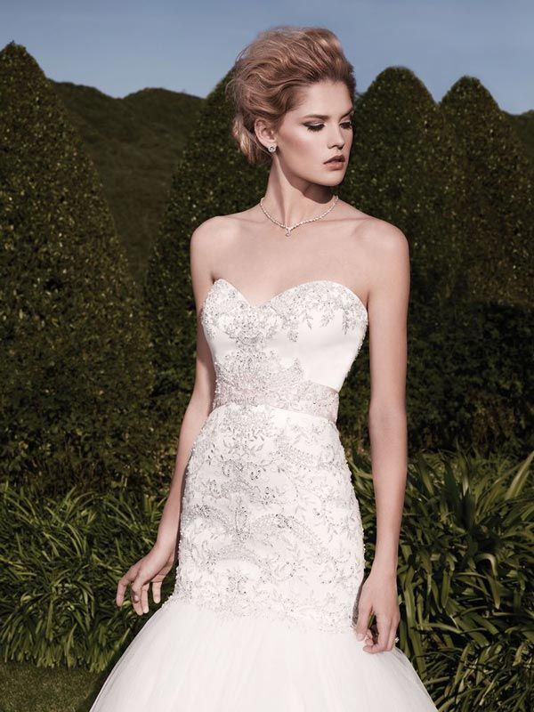 Woman in a white strapless lace gown standing outdoors in front of a hedge backdrop