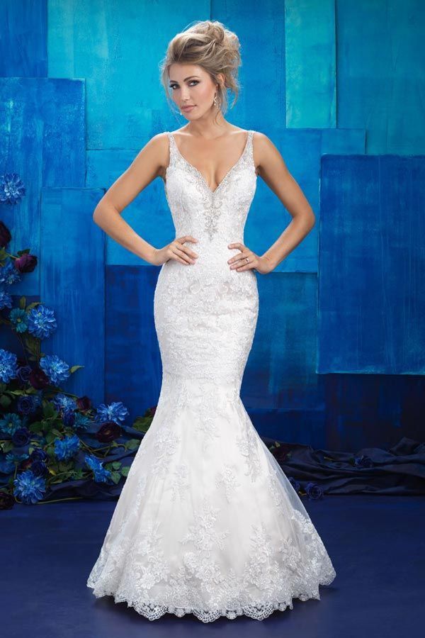 Woman in a white lace wedding dress standing against a blue backdrop, hands on hips.