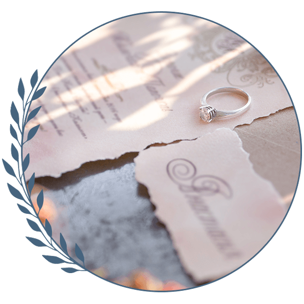 Wedding invitation with silver ring on torn paper, soft pink tones, framed by leafy wreath