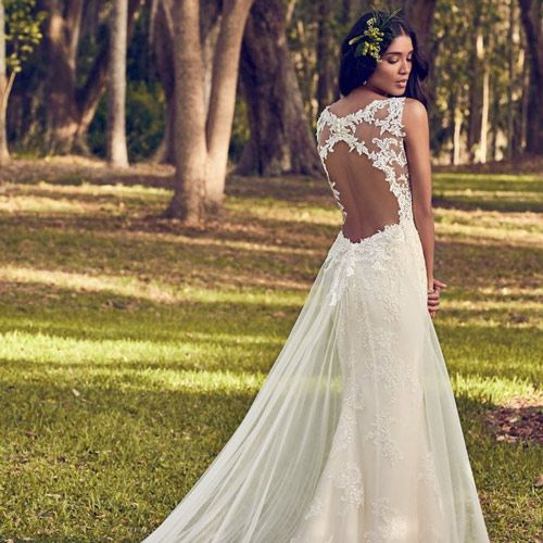 Woman in a white lace gown standing in a wooded park, seen from behind.