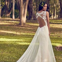 Woman in a white lace gown standing in a wooded park, seen from behind.