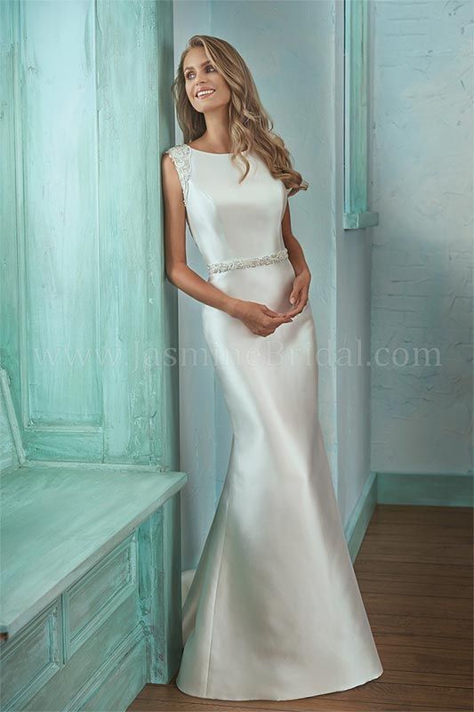 Woman in a white satin gown standing in a pastel turquoise room, leaning against a wall.